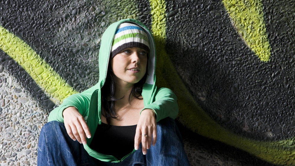 Bilde av en jente med grønn hettegenser som sitter foran en vegg med graffiti.